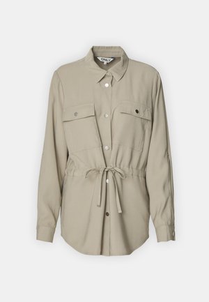 Světle béžová košile na knoflíky s límečkem, dvěma náprsními kapsami a páskem v pase. Má dlouhé rukávy a stříbrné knoflíkové detaily.