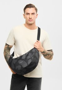 Čierna crossbody taška s nepravidelným tvarom, textúrou látky a nastaviteľným popruhom. Logo viditeľné na prednej strane.