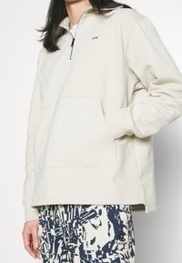 Světle béžový pulovr se čtvrtinovým zipem, který má přední kapsu a žebrované manžety. Materiál působí měkce a má hladkou texturu.