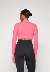 Pink cropped long-sleeve top with a high collar, fitted design, and flat seams. Paired with high-waisted black leggings, smooth texture.