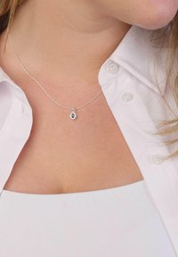 Collier en argent avec un pendentif floral présentant une pierre centrale bleue et des accents en argent, porté sur une chemise blanche.