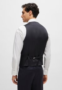 Schwarze Weste mit einem strukturierten Muster, verstellbarem Rückengurt, getragen über einem weißen Hemd. Klare Linien und taillierte Passform, die die Silhouette betont.