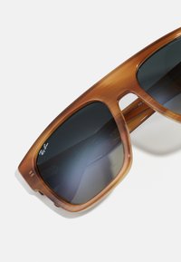 Ochelari de soare din plastic cu un gradient de culoare maronie, cu lentile fumurii negre, design unghiular și detaliu cu logo argintiu pe lentila stângă.