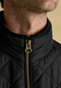 Czarna pikowana kurtka z wysokim kołnierzem, z złotym zamkiem błyskawicznym z logo "JOULES", widocznymi szwami i gładką teksturą.