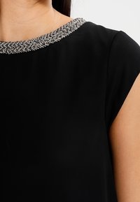 Black dress featuring a round neckline adorned with a woven silver beaded trim. Smooth texture with short sleeves.