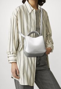 Osoba ubrana w luźną beżowo-czarną koszulę w paski oraz szare dżinsy, niosąca małą srebrną torebkę na pasku na przodzie.