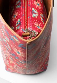 Bolso con un exterior de tela rojo con un patrón floral, que presenta un cierre metálico y una lengüeta de cuero, exhibiendo diseños florales en azul y amarillo.