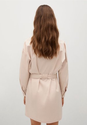 Femme aux cheveux longs et ondulés bruns porte un manteau beige clair ceinturé, debout devant un fond blanc uni, dos à la caméra.