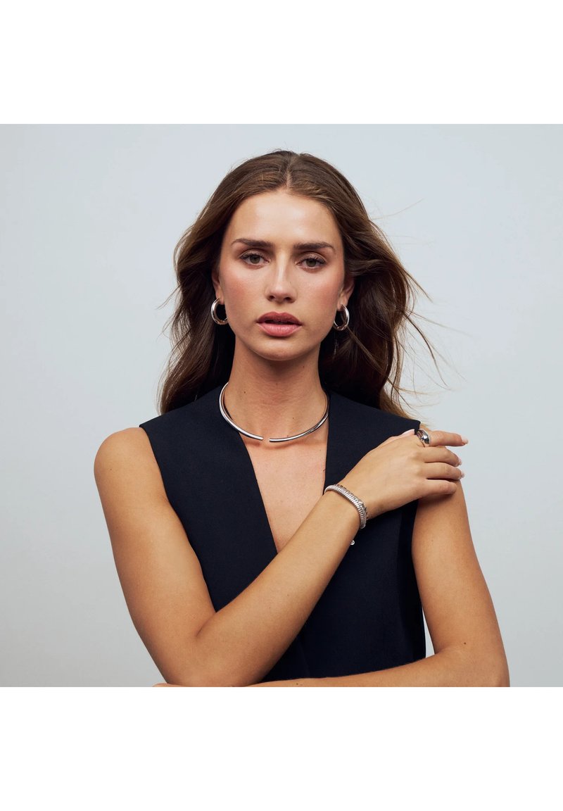 Smycken i silver som består av en stilren choker, hoopörhängen, ett texturerat armband och en ring, mot en enkel bakgrund.