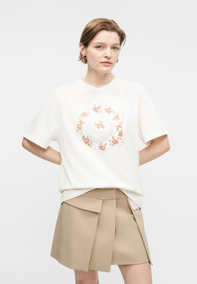 Kvinne med kort hår iført en hvit T-skjorte med blomstermotiv i en sirkel og beige plissettert miniskjørt, står med hendene bak ryggen.