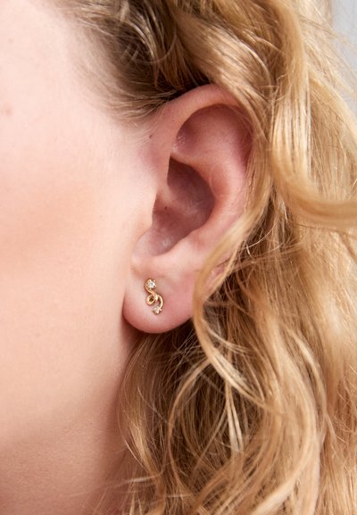 Gold-toned earrings with a swirl design, featuring a small gem accent. The earrings are positioned in the earlobe, next to loose, wavy hair.