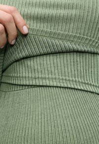 Χέρι σηκώνει το τελείωμα ενός πράσινου πλεκτού ενδύματος με ριμπ υφή, δείχνοντας τις λεπτομερείς ραφές και την υφή του υφάσματος.
