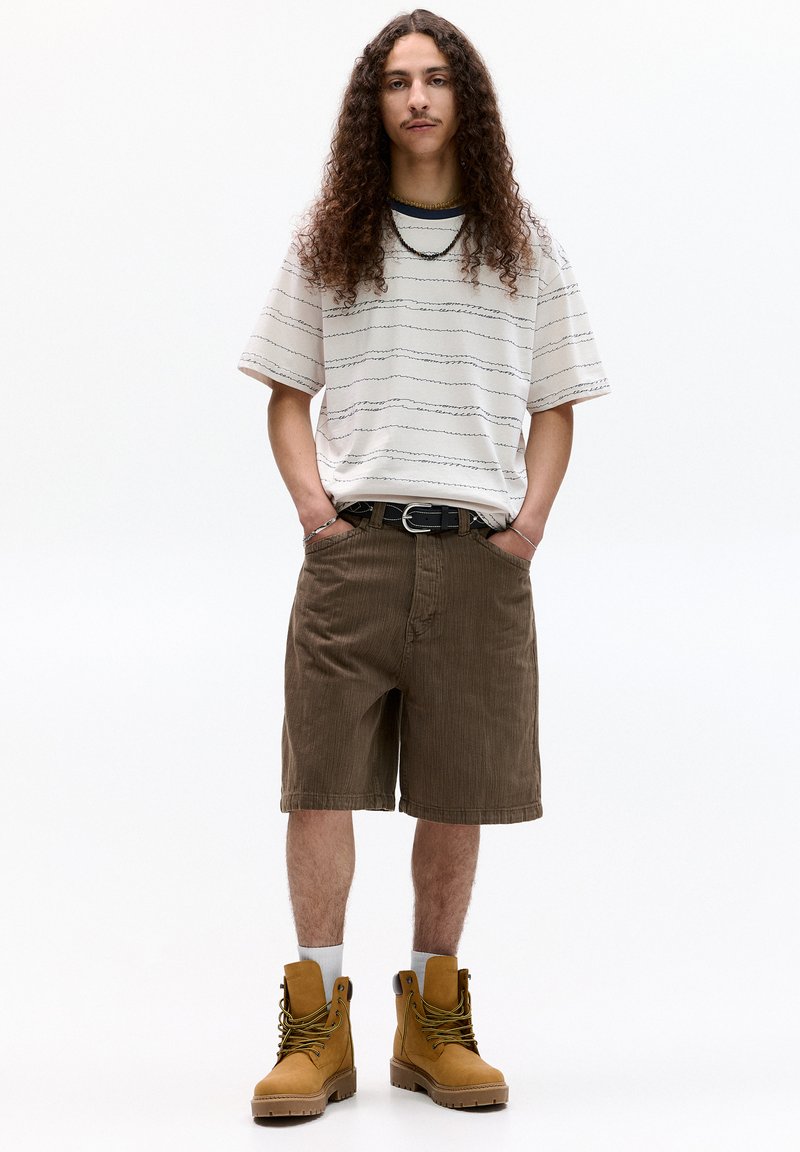 Khaki corduroy shorts, hvid og sort stribet t-shirt, brune snørebåndsstøvler. Modellen har langt krøllet hår og hænderne i lommerne. Enkel design.