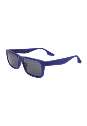 Occhiali da sole rettangolari con montatura blu, lenti scure leggermente curve e logo Converse sull'interno dell'asta.