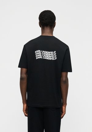 Homme portant un t-shirt noir avec "KARL LAGERFELD" imprimé quatre fois en blanc sur le haut du dos, debout devant un fond uni.