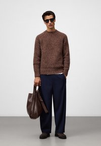 Brązowy sweter z teksturowanym wzorem, granatowe szerokie spodnie, ciemnobrązowa skórzana torba, czarne okulary przeciwsłoneczne, stojąca na szarym podłodze na tle białej ściany.