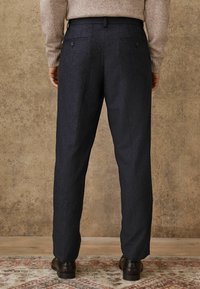 Pantalons ajustés bleu marine avec une finition texturée, dotés de deux poches arrière et d'accents en métal sur les boutons. Les jambes se rétrécissent légèrement vers l'ourlet.