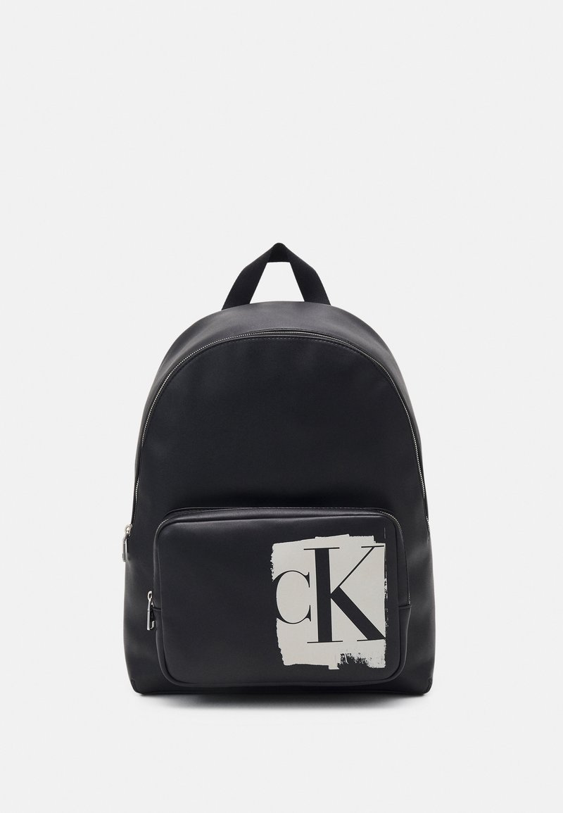 Sac à dos noir avec une poche avant zippée présentant un patch blanc et un grand logo noir "cK", et une poignée de transport supérieure.