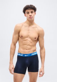 Ευθυτενής νέος άνδρας με σγουρά μαλλιά, που φοράει μαύρα boxer briefs Nike με μπλε ζώνη στη μέση, στέκεται μπροστά σε απλό ανοιχτό φόντο.