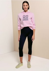 Femme portant un sweat à capuche rose avec le texte « sea », des leggings noirs courts et des baskets beiges, debout devant un fond bicolore beige et blanc.