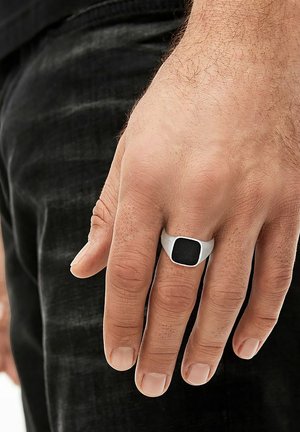 Silberner metallischer Ring mit einem schwarzen quadratischen Mittelpunkt, getragen an einer Hand mit kurzen, hellen Nägeln. Der Finger ist teilweise gebogen.