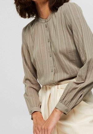Kvinde iført en beige skjorte med lange ærmer og knapper med subtile lodrette striber og off-white højtaljede bukser bundet i taljen.