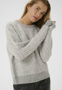 Kvinde iført en grå og hvid zigzag-mønstret sweater med ribbede manchetter og kant, kombineret med sorte læderbukser, poserende mod en hvid baggrund.