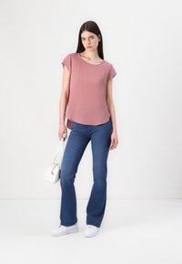 Top rosa chiaro a maniche corte con scollatura arrotondata, abbinato a jeans a zampa d'elefante blu chiaro e sneakers bianche, mentre tiene una piccola borsa bianca.