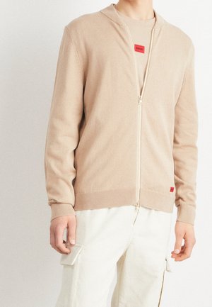 Man draagt een beige cardigan met rits over een beige shirt met rood logo, gecombineerd met witte cargobroek, staand tegen een effen achtergrond.