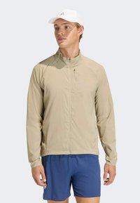 Ανοιχτό μπεζ ελαφρύ μπουφάν με υψηλό γιακά, με φερμουάρ μπροστά και τσέπες με φερμουάρ; φορεμένο πάνω από μπλε �σορτς; λείος υφή, αθλητικός σχεδιασμός.