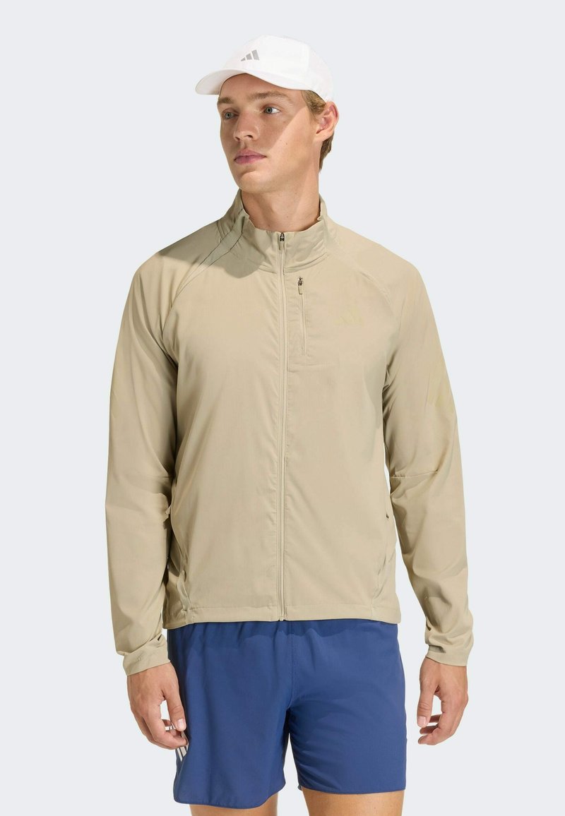 Ανοιχτό μπεζ ελαφρύ μπουφάν με υψηλό γιακά, με φερμουάρ μπροστά και τσέπες με φερμουάρ; φορεμένο πάνω από μπλε σορτς; λείος υφή, αθλητικός σχεδιασμός.