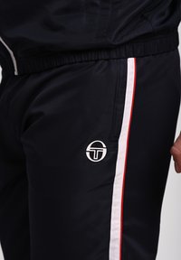 Námořnické sportovní kalhoty vyrobené z texturované tkaniny, s bílými a červenými bočními pruhy a kulatým logem na stehně. Elastický pas.