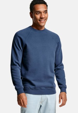 Glimlachende man met een blauw crewneck sweatshirt en lichtblauwe spijkerbroek, staand tegen een effen grijze achtergrond.