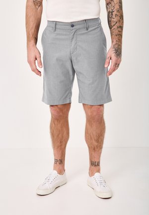 Male wearing light gray knee-length shorts and white sneakers, standing against a plain background with visible arm and leg tattoos.
