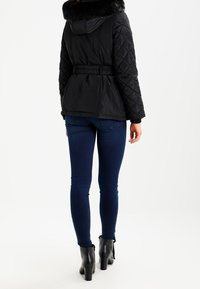 Mustat kiiltäväpintaiset takki, jossa on keinoturkiskaulus, vyötärö on kiristetty ja elastisia yksityiskohtia, yhdistettyinä sinisiin istuviin farkkuihin ja mustiin nilkkureihin.