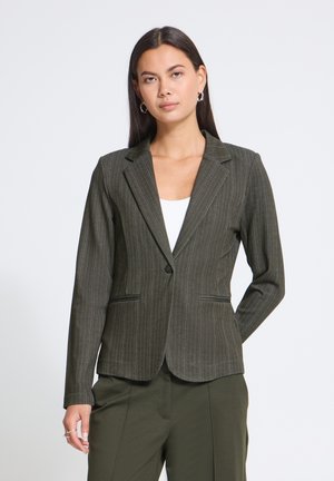 Femme portant un blazer à fines rayures vert olive foncé sur un haut blanc et un pantalon olive assorti, debout avec une main derrière le dos.