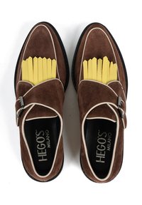 Brązowe zamszowe mokasyny z paskiem ze srebrnym hardwarem, kontrastującym białym obszyciem i żółtymi akcentami przypominającymi frytki na obszarze palców.