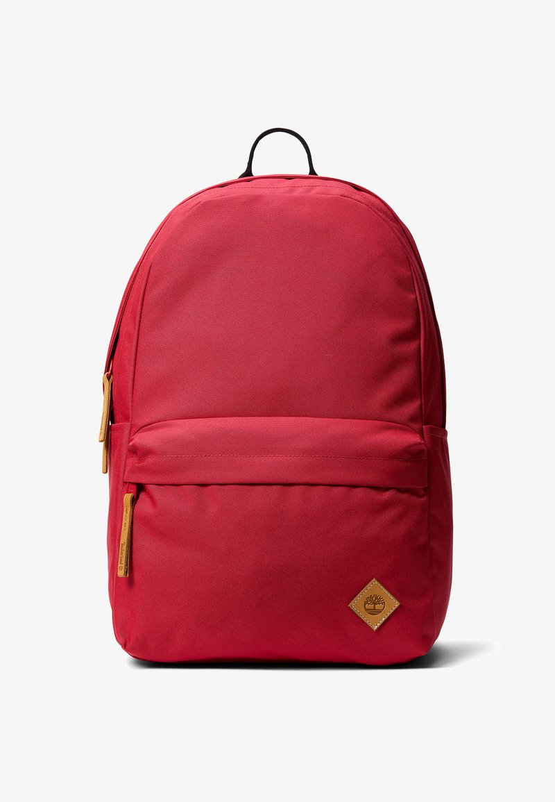 Sac à dos en tissu rouge avec une poignée supérieure, une poche zippée à l'avant, et un patch logo en cuir. Texture lisse avec une forme épurée et arrondie.