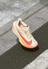 Beige sportschoen met een bovenwerk van gaas, grote roze swoosh, vergezeld van gele en oranje details op de zool, met een gestructureerd patroon.