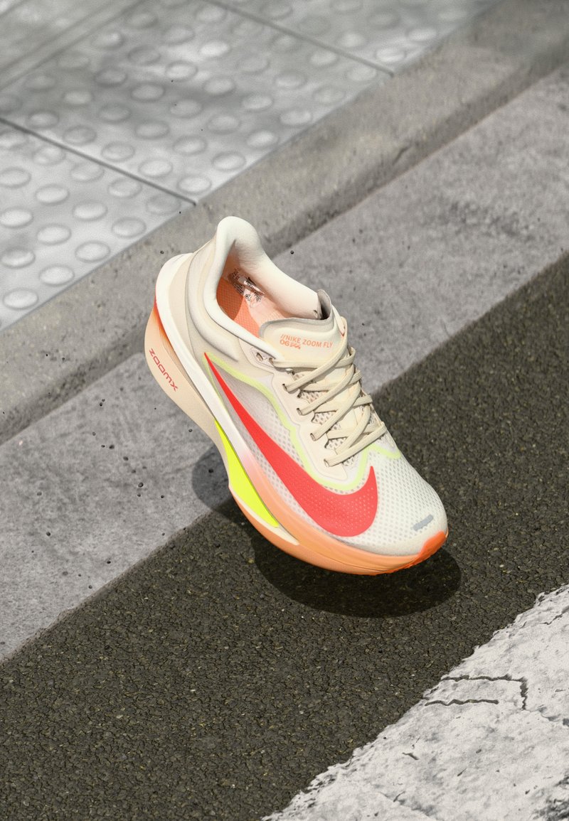 Beige sportschoen met een bovenwerk van gaas, grote roze swoosh, vergezeld van gele en oranje details op de zool, met een gestructureerd patroon.