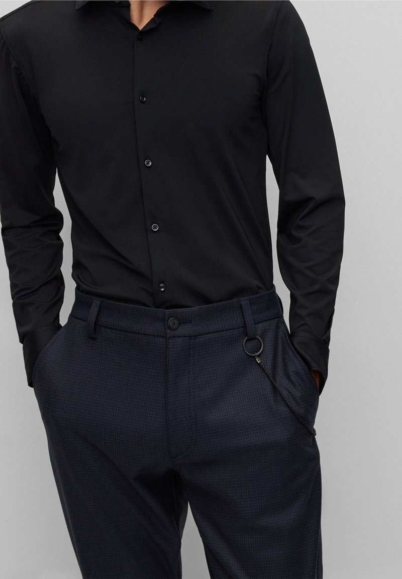 Homme portant une chemise noire cintrée à boutons rentrée dans un pantalon à carreaux foncé, les mains dans les poches, sur un fond uni.