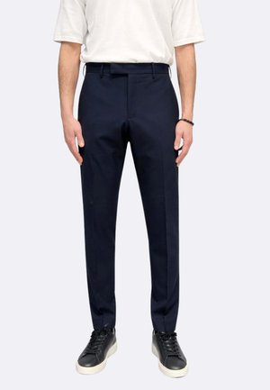 Uomo che indossa pantaloni slim fit blu navy, t-shirt bianca, sneakers nere e un braccialetto nero con perline, in piedi con le mani rilassate ai lati.