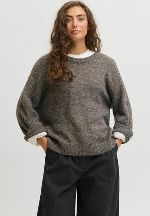 Donna con lunghi capelli ricci che indossa un maglione grigio ampio sopra una camicia bianca e pantaloni neri a gamba larga, mani nelle tasche.
