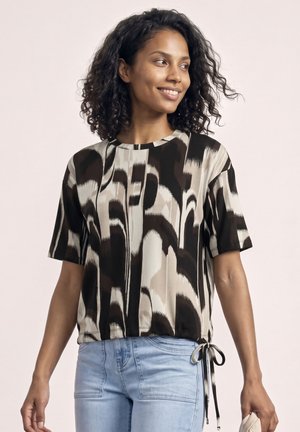 Femme aux cheveux noirs bouclés souriante, portant un chemisier à manches courtes à motif abstrait noir et beige et un jean bleu clair, tenant un objet beige.