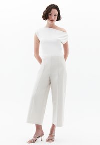 Bílý top na jedno rameno v kombinaci s smetanovými kalhotami s širokými nohavicemi. Oblečení je vyrobeno z hladkých tkanin a má jemné skladby. Doplňují ho sandály na vysokém podpatku.