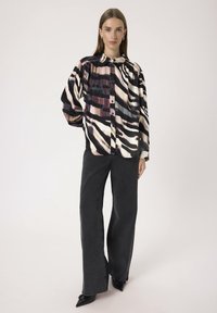 Kvinde står iført en langærmet skjorte med abstrakte mønstre i sort, hvid og lilla, sorte vide jeans og sorte spidse sko.