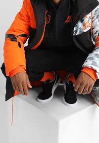 Giubbotto gonfiabile nero e arancione con dettagli in camouflage, sneakers nere e calzini arancioni. Modello accosciato su un piedistallo bianco.
