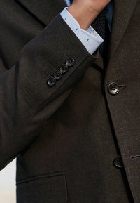 Sacou texturat de culoare închisă, cu patru butoni negri la manșon și cu un manșon de cămașă rayat de culoare albastru deschis care iese în evidență.