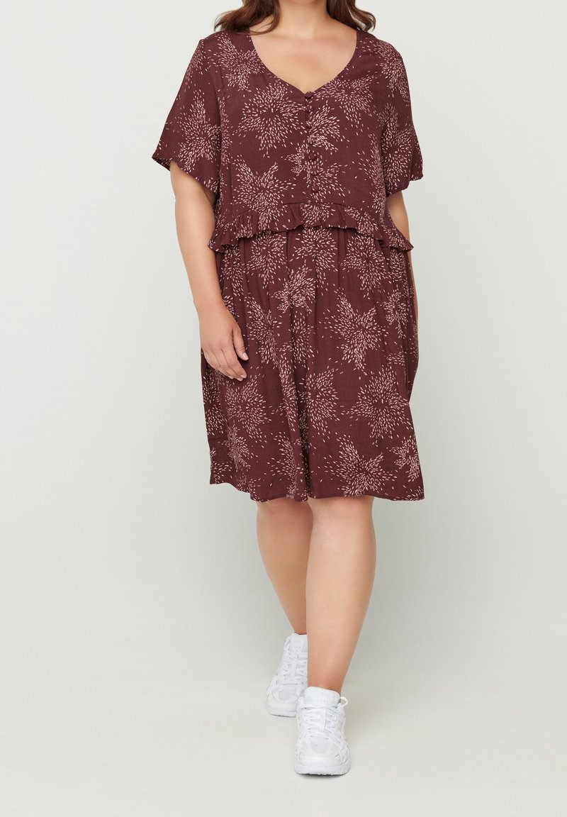 Robe bordeaux avec un décolleté en V, des manches courtes et une volée à la taille. Présente un motif floral blanc et est associée à des baskets blanches.