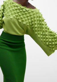 Maglione verde con un motivo testurizzato e a punte sulle maniche, abbinato a una gonna verde aderente. Tessuto a maglia morbido, vestibilità ampia sulle spalle.